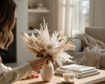 Op de afbeelding: Een neutraal bloemstuk in een crèmekleurige vaas. Het boeket bevat gedroogd pampasgras, tarwe en een witte roos. De vaas staat op een licht houten salontafel, met een kopje thee en boeken in de buurt. De scène baadt in zacht, natuurlijk licht.