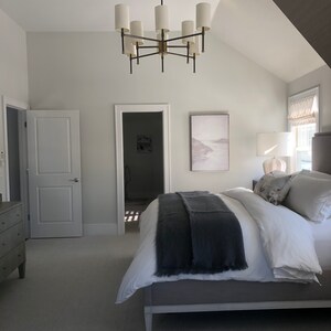 May include: A bedroom with a large bed with a gray blanket and white bedding. There is a large chandelier with white shades hanging from the ceiling. There is a dresser on the left side of the bed and a painting on the wall behind the bed.
