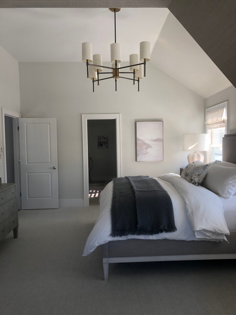 May include: A bedroom with a large bed with a gray blanket and white bedding. There is a large chandelier with white shades hanging from the ceiling. There is a dresser on the left side of the bed and a painting on the wall behind the bed.
