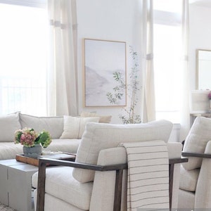 May include: A white living room with a white sofa and two white armchairs. The armchairs have dark wood frames and one has a white and gray striped throw blanket draped over it. There is a small table in front of the sofa with a vase of pink flowers on it. A framed print hangs on the wall behind the sofa.