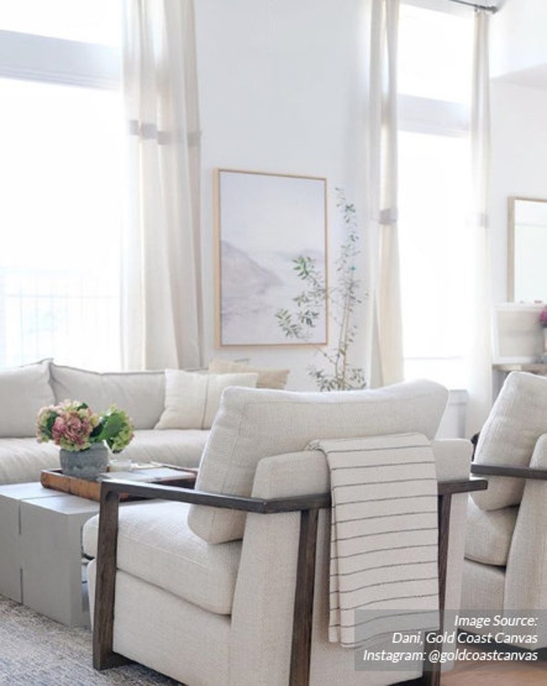 May include: A white living room with a white sofa and two white armchairs. The armchairs have dark wood frames and one has a white and gray striped throw blanket draped over it. There is a small table in front of the sofa with a vase of pink flowers on it. A framed print hangs on the wall behind the sofa.