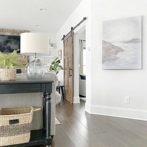 May include: A black console table with a woven basket on the bottom shelf. The table is in a living room with a sliding barn door, a white lamp, and a painting on the wall.
