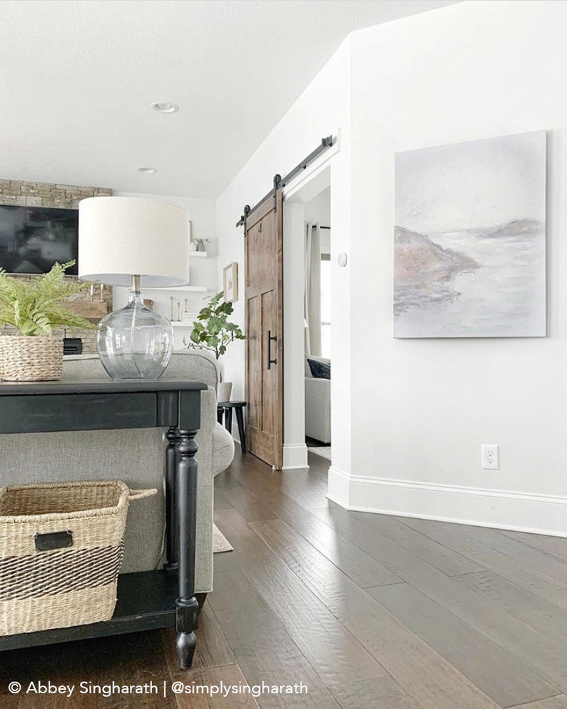 May include: A black console table with a woven basket on the bottom shelf. The table is in a living room with a sliding barn door, a white lamp, and a painting on the wall.