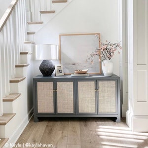 May include: A grey cabinet with four doors and woven cane panels. The cabinet has brass knobs and sits on a wooden floor. A white lamp with a black base and a white vase with pink flowers are on top of the cabinet. A framed painting hangs on the wall above the cabinet.