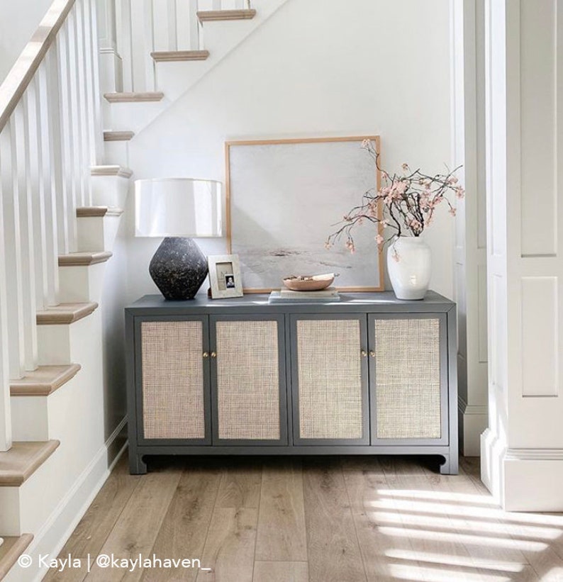 May include: A grey cabinet with four doors and woven cane panels. The cabinet has brass knobs and sits on a wooden floor. A white lamp with a black base and a white vase with pink flowers are on top of the cabinet. A framed painting hangs on the wall above the cabinet.