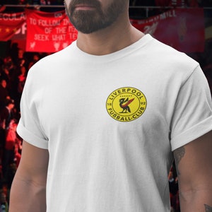 May include: White t-shirt with a yellow circle logo featuring a red bird and the text "LIVERPOOL FOOTBALL CLUB 1892".