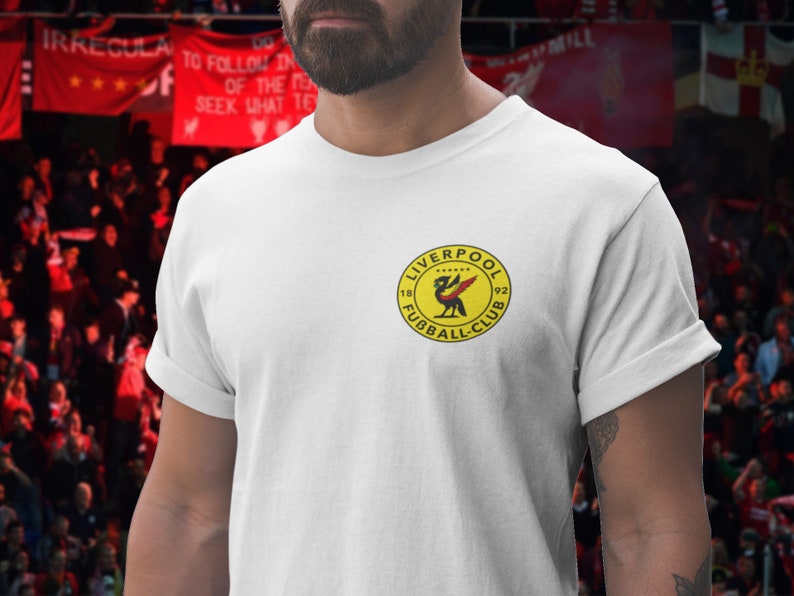 May include: White t-shirt with a yellow circle logo featuring a red bird and the text "LIVERPOOL FOOTBALL CLUB 1892".