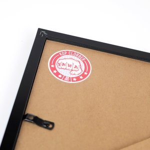 May include: Black picture frame with a red and white sticker that says "Kop Clobber" with a fist graphic and the year 2017.