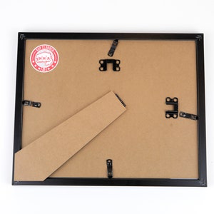 May include: The back of a black picture frame with a brown cardboard backing. The frame has metal hanging hardware and a red and white sticker that says "Top Clobber".