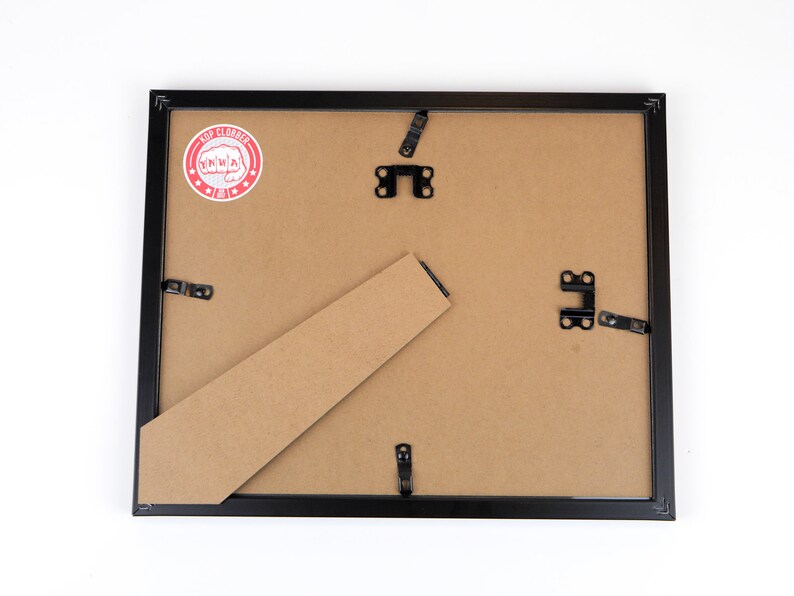 May include: The back of a black picture frame with a brown cardboard backing. The frame has metal hanging hardware and a red and white sticker that says "Top Clobber".