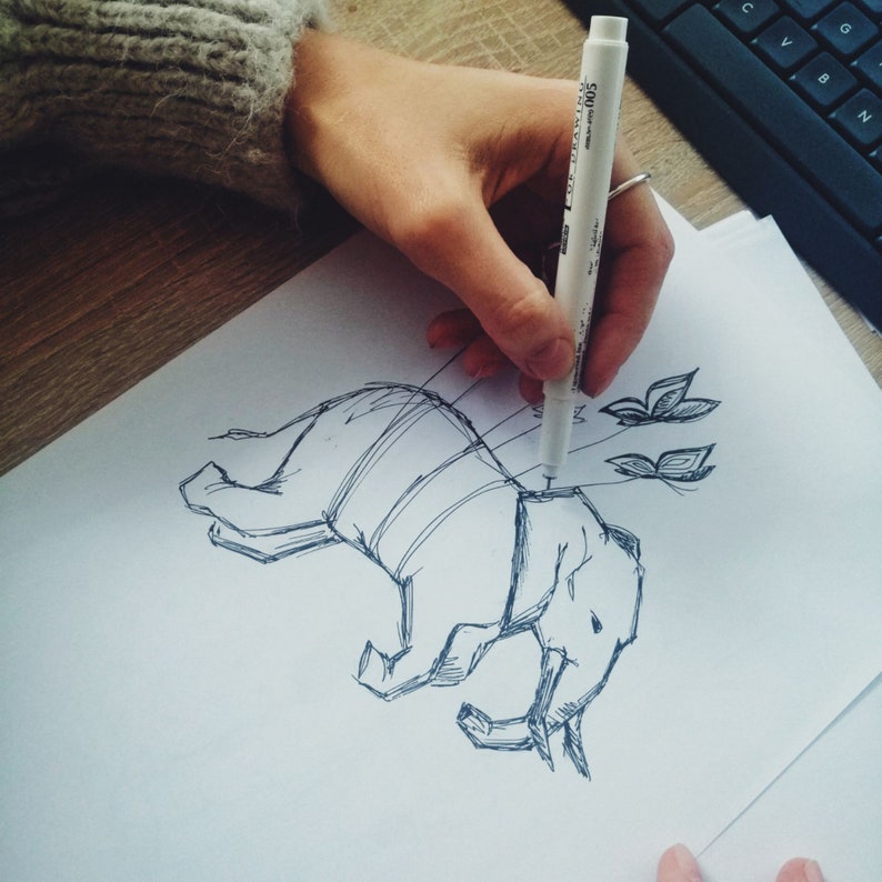 May include: A hand draws a black and white sketch of an elephant with butterflies on a white sheet of paper. The elephant is drawn with a striped pattern and is in a playful pose.