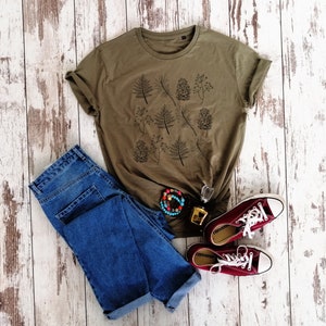 May include: A green t-shirt with a black and white print of leaves and pine cones. The t-shirt is paired with blue jeans and maroon converse shoes.
