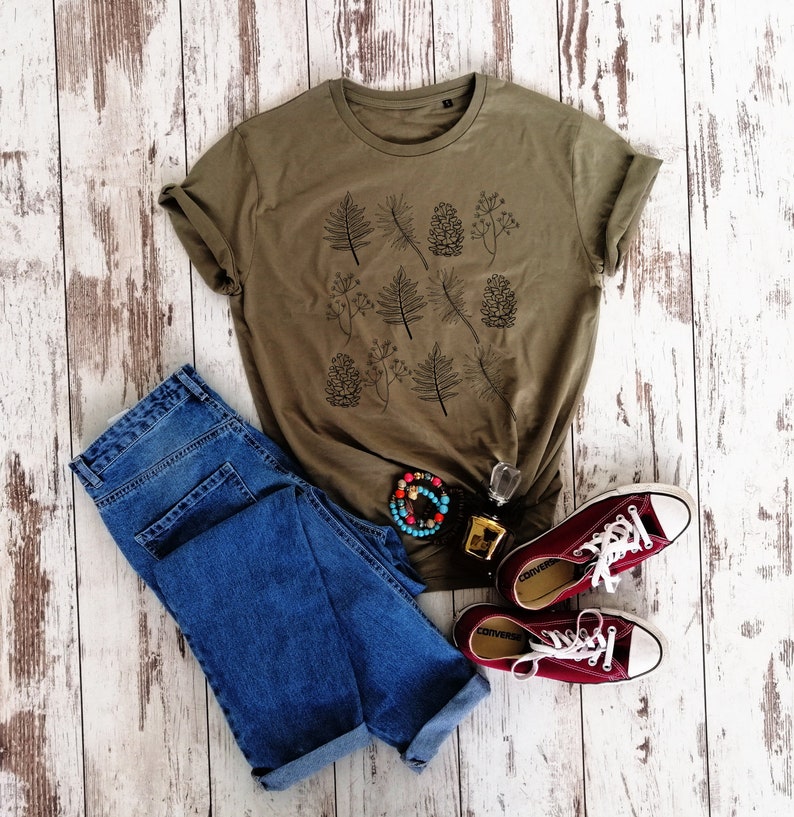 May include: A green t-shirt with a black and white print of leaves and pine cones. The t-shirt is paired with blue jeans and maroon converse shoes.