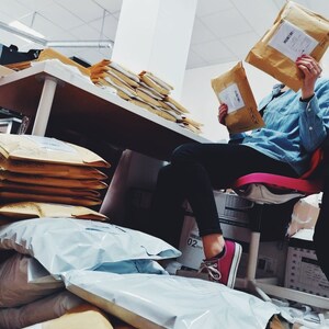 May include: A person is sitting on a chair surrounded by stacks of brown envelopes and white padded envelopes. The person is wearing a blue denim shirt and pink sneakers. The person is holding a brown envelope in their right hand.