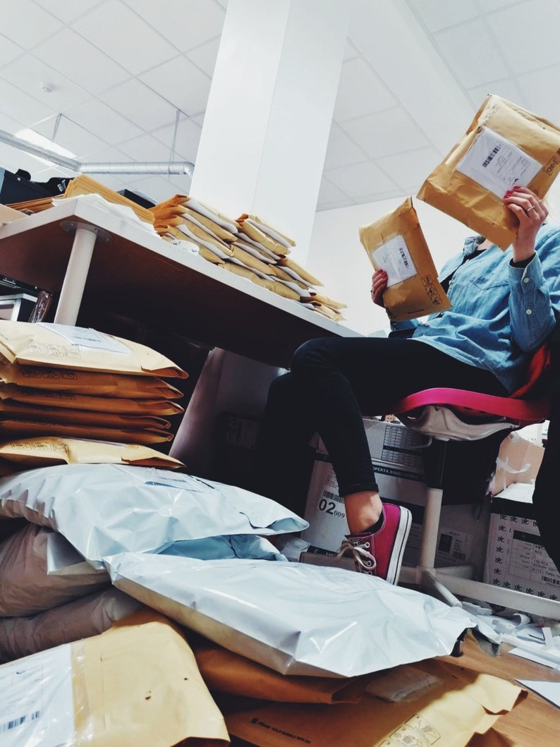 May include: A person is sitting on a chair surrounded by stacks of brown envelopes and white padded envelopes. The person is wearing a blue denim shirt and pink sneakers. The person is holding a brown envelope in their right hand.