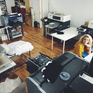 May include: A woman sits at a computer in a workshop with several large printing machines. The machines are used for printing on fabric and other materials.