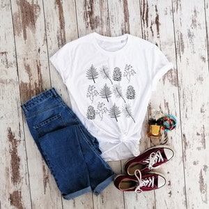 May include: A white t-shirt with a black and white print of pine cones and leaves. The shirt is styled with blue jeans and maroon sneakers.