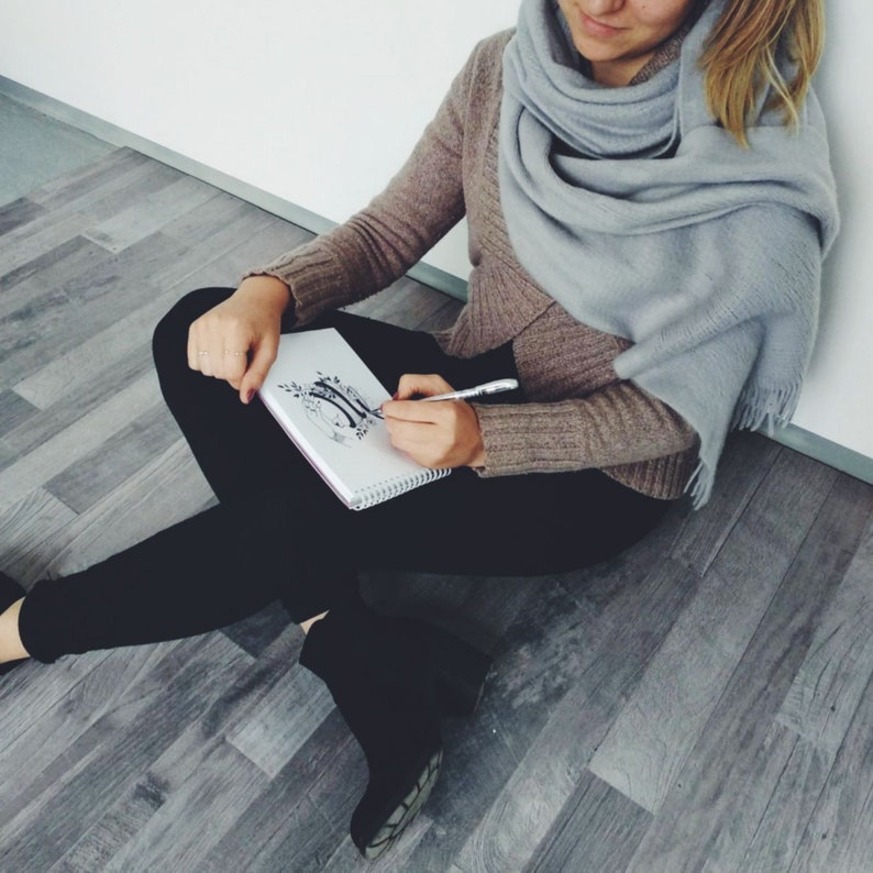 May include: A person sitting on the floor with a sketchbook open on their lap. They are holding a pen and drawing in the sketchbook. The sketchbook has a floral design on the cover.