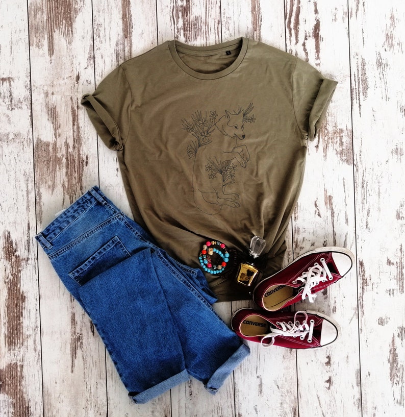 May include: A green t-shirt with a line drawing of a wolf surrounded by flowers. The t-shirt is laying on a white wooden surface with a pair of blue jeans and red sneakers.