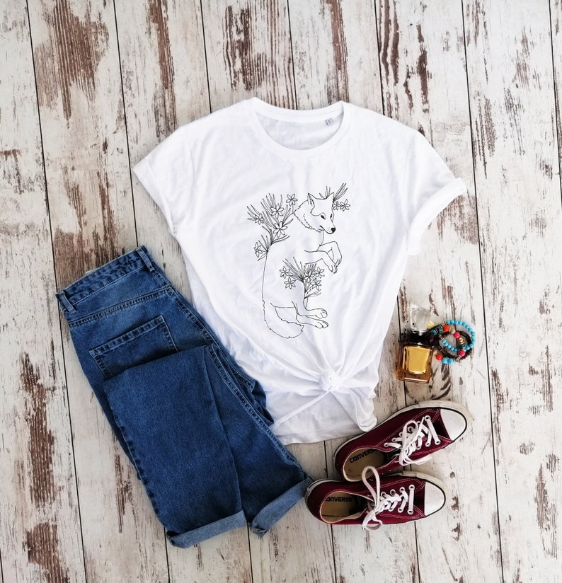May include: White t-shirt with a black line drawing of a wolf surrounded by flowers. The shirt is laid out on a wooden surface with a pair of blue jeans, a bottle of perfume, and a bracelet.
