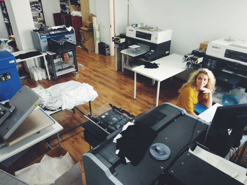 May include: A woman sits at a computer in a workshop with several large printing machines. The machines are used for printing on fabric and other materials.