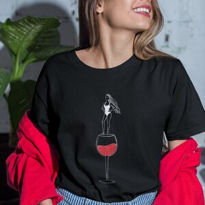 May include: Black t-shirt with a graphic of a woman standing in a wine glass. The woman is wearing a swimsuit and has long hair. The wine glass is filled with red wine.