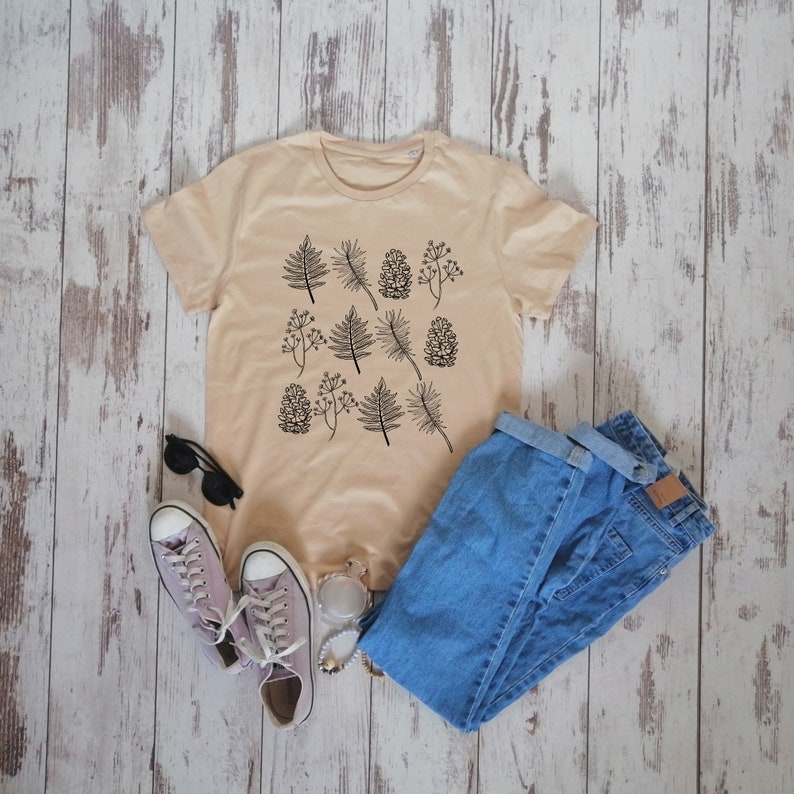 May include: A beige t-shirt with a black and white print of pine needles and pine cones. The shirt is laid flat on a light wood surface with a pair of light purple sneakers, sunglasses, and a pair of blue jeans.