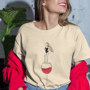 May include: A light beige t-shirt with a graphic of a woman standing on top of a glass of red wine. The woman is wearing a black swimsuit and has long hair.