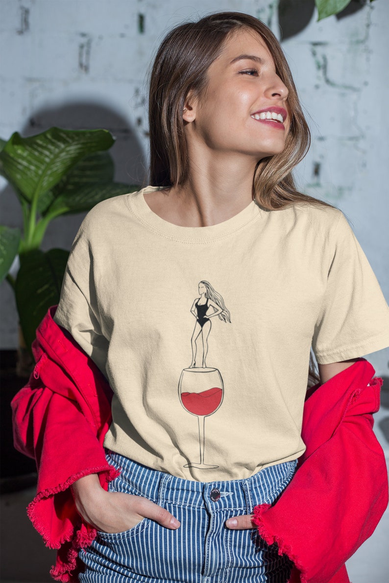 May include: A light beige t-shirt with a graphic of a woman standing on top of a glass of red wine. The woman is wearing a black swimsuit and has long hair.