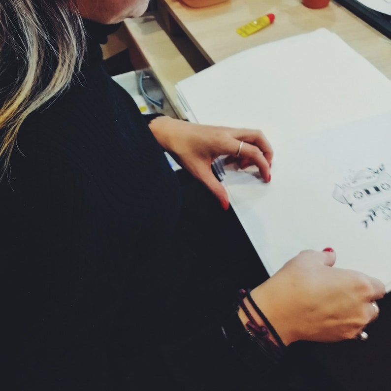 May include: A person is drawing on a white sheet of paper. The paper is on a table and the person is sitting at the table. The person is wearing a black shirt and has long blonde hair. The person is using a black pen to draw.