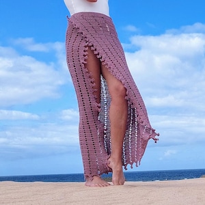 Puede incluir: Una falda de crochet malva con una abertura alta, usada con un top blanco. La falda tiene un patrón texturizado y un dobladillo con flecos. El fondo presenta un cielo azul con nubes y una playa arenosa.