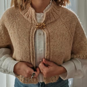 以下が含まれることがあります： ベージュ色のニットボレロと金色の花のブローチ。半袖セーターは、白いボタンダウンシャツとブルージーンズの上に着用されています。セーターはハイネックで、ブローチで留められています。