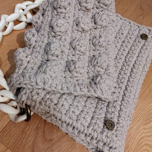 Puede incluir: Un bolso de mano de ganchillo gris claro con una correa de cadena blanca. El bolso presenta un patrón texturizado con detalles redondeados en relieve. La solapa se asegura con un cierre de botón a presión de color bronce. El bolso está sobre una superficie de madera.