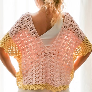 Puede incluir: Top de ganchillo rosa pastel y amarillo con diseño de cuello en V. El top tiene un patrón floral y un ribete amarillo. La prenda se lleva sobre una camiseta blanca. El ganchillo es probablemente hecho a mano.