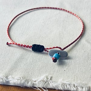 May include: A red and white string bracelet with a black bead and a silver toggle clasp. The bracelet has a blue bead on the toggle clasp.