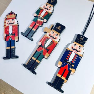 May include: Four wooden nutcracker ornaments with different designs. The ornaments are painted in red, blue, green, and black with gold accents. The nutcrackers are wearing hats and have mustaches.