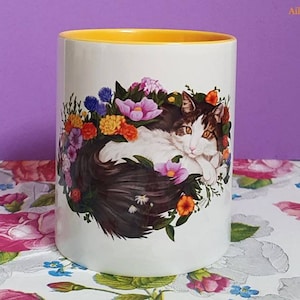 Puede incluir: Taza de cerámica blanca con borde amarillo. La taza presenta una ilustración colorida de un gato rodeado de flores.