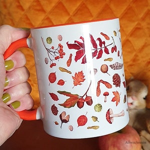 Könnte beinhalten: Eine weiße Keramiktasse mit orangefarbenem Rand und Henkel. Die Tasse zeigt ein Muster aus Herbstblättern, Eicheln, Pilzen und anderen Waldelementen in Rot-, Orange- und Brauntönen.