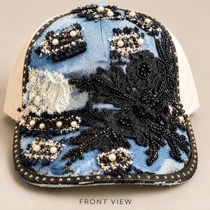 May include: A light blue denim baseball cap featuring a large black lace floral appliqué with beadwork, cream-colored pearls, and frayed fabric patches. The brim is edged with a black trim accented by small gold studs, and the side panels are beige.