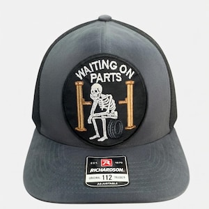 Può includere: Un cappellino trucker grigio antracite con una toppa nera circolare che mostra uno scheletro bianco ricamato seduto su uno pneumatico tra due pilastri dorati. Il testo WAITING ON PARTS è arcuato sopra lo scheletro. Un adesivo Richardson 112 è sulla visiera.