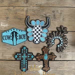 May include: Five embroidered patches featuring a light blue hexagonal patch with the text COWBOY, a black and white checkered bull skull with light blue accents, a horseshoe shape with brown floral patterns, and two crosses in black, brown, and blue.