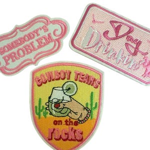 Puede incluir: Tres parches bordados. Uno es de forma festoneada rosa con el texto "SOMEBODY'S PROBLEM". Otro es un rectángulo rosa con el texto "Day Drinkin". El tercero es un parche en forma de escudo con una mano sosteniendo una bebida, cactus y "COWBOY TEARS on the rocks".