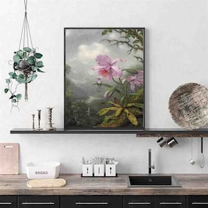 Puede incluir: Una impresión botánica enmarcada que presenta orquídeas rosas en un paisaje exuberante y verde. La obra de arte se exhibe sobre una encimera de cocina con gabinetes negros, un fregadero y una panera. También se ven una planta colgante y objetos decorativos.