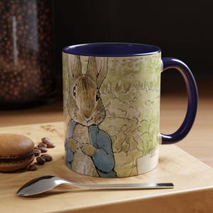 Puede incluir: Una taza de cerámica azul y blanca con una imagen de Peter Rabbit. La taza tiene un borde y un asa azules.