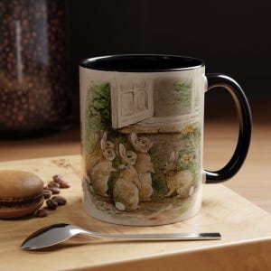 Puede incluir: Una taza de cerámica blanca con un borde y un asa negros. La taza presenta una ilustración colorida de cinco conejos de pie frente a una puerta. Los conejos son marrones y blancos con narices y bigotes negros.