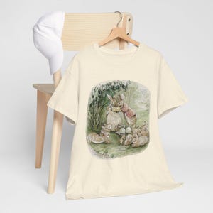 May include: A beige t-shirt featuring a print of Peter Rabbit and his family. The print depicts Peter Rabbit holding a basket of carrots, surrounded by other rabbits.