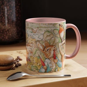 Puede incluir: Una taza de cerámica con interior y asa rosas, con una ilustración caprichosa de un conejo con chaqueta azul comiendo una zanahoria, con un pájaro posado en una rama. El fondo representa una escena de jardín. Una cuchara y un pastel están sobre una superficie de madera.