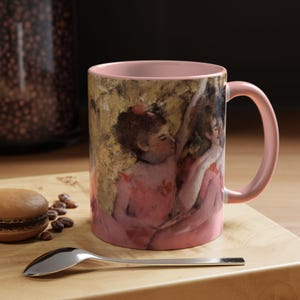 Puede incluir: Una taza de cerámica rosa con asa presenta una pintura de dos bailarinas con vestidos rosas. Las bailarinas están en un abrazo cercano, con la cabeza inclinada la una hacia la otra.