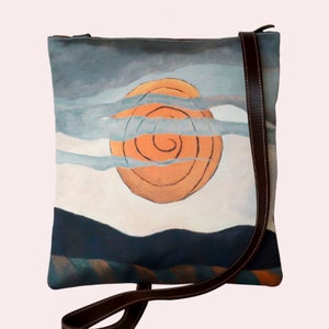 Puede incluir: Un bolso bandolera de lona con una correa de cuero marrón. El bolso presenta un diseño pintado de un sol naranja giratorio sobre un cielo azul y blanco con una cordillera azul oscuro en primer plano.
