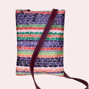 Puede incluir: Un bolso bandolera de rayas coloridas con un patrón de acuarela morado, rosa, verde y naranja. El bolso tiene una correa ajustable de color burdeos.